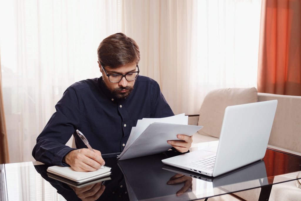 Entrepreneur in eyeglasses works with a laptop and keeps a document in a home office. Man holding paper documents, chatting online with clients on laptop at workplace.