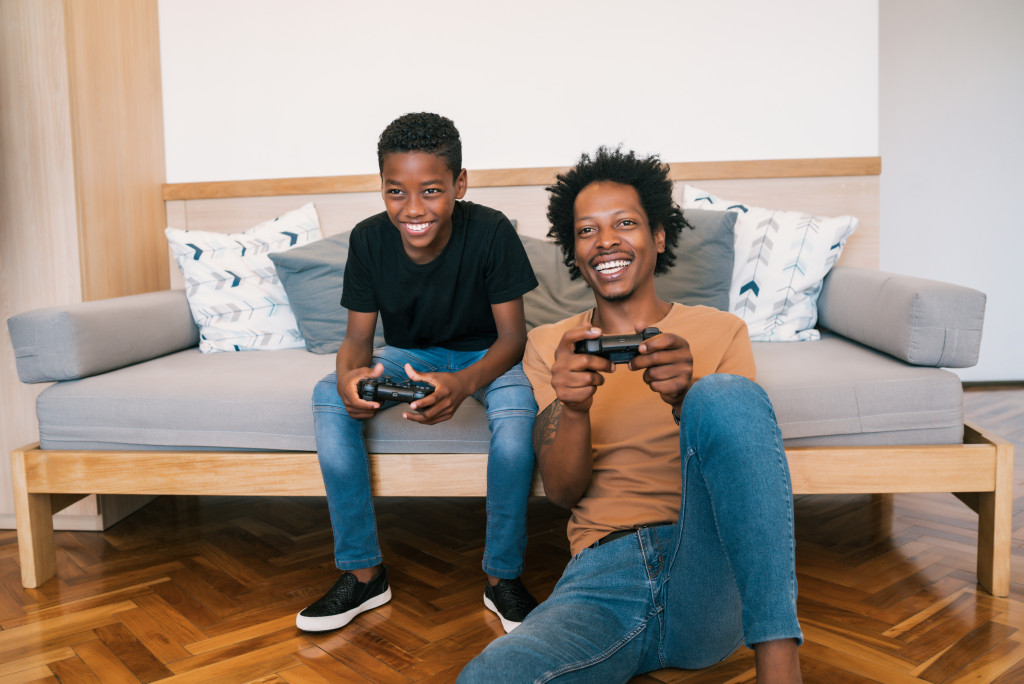 Portrait of happy African American father and son sitting in sofa couch and playing console video games together at home. Family and technology concept.