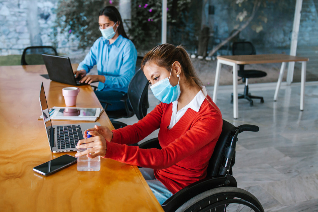 A woman in a wheelchair wearing her mask, and another woman beside her typing on a laptop at their workplace.